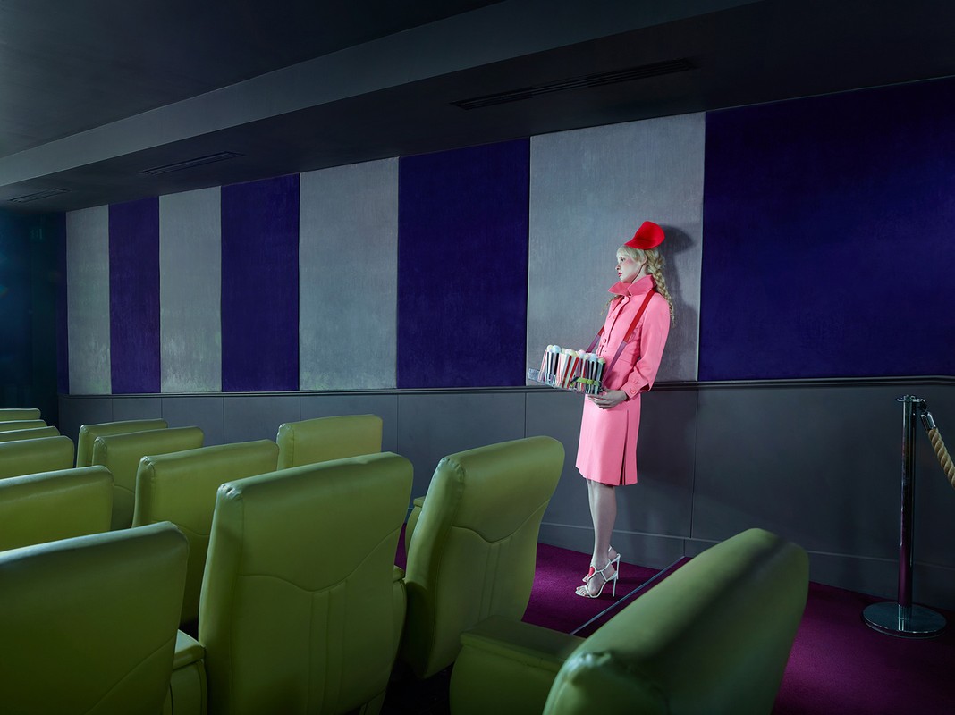 Fashion shot in a vintage movie theater, featuring a woman in a pink coat and red hat holding a snack tray against striped wall.