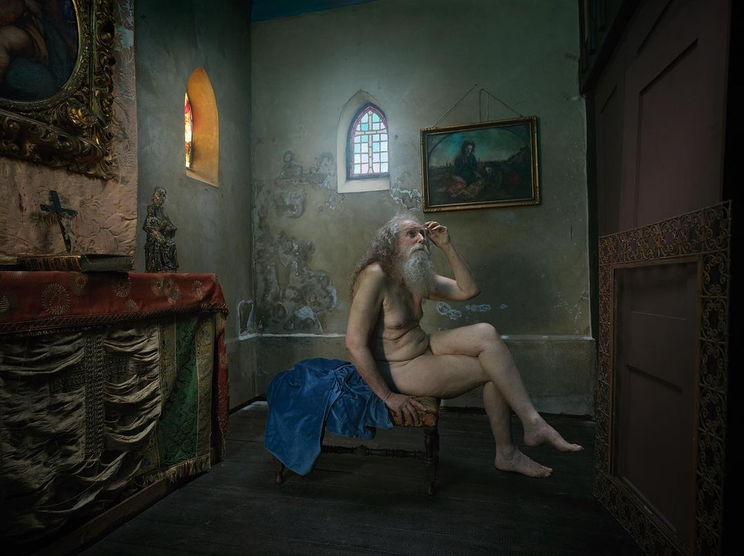 Nude man sits on a chair in an old church with art and stained glass visible in the background, creating a fine art photography scene.