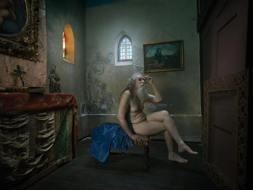 Nude man sits on a chair in an old church with art and stained glass visible in the background, creating a fine art photography scene.