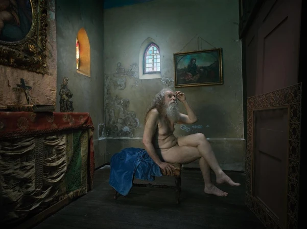Nude man sits on a chair in an old church with art and stained glass visible in the background, creating a fine art photography scene.