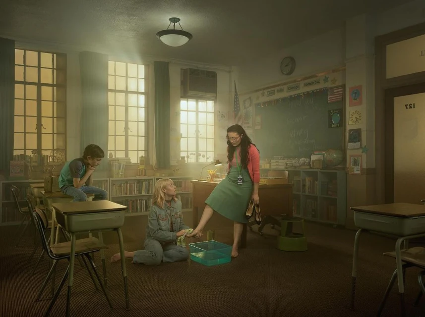 In a classroom, a woman stands with her foot in a tub as another woman kneels nearby, while a young boy sits on a desk in the background.