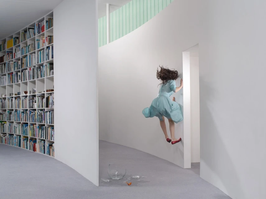 A woman in a blue dress appears to float in a minimalist interior with white walls, a bookcase, and a broken vase in the foreground.
