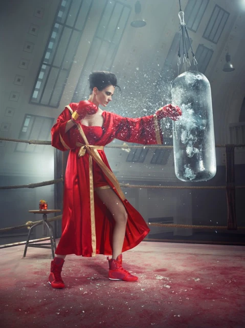 Female boxer in red robe and boxing gloves punches a clear punching bag in boxing ring with dramatic lighting and a stool with a beverage.