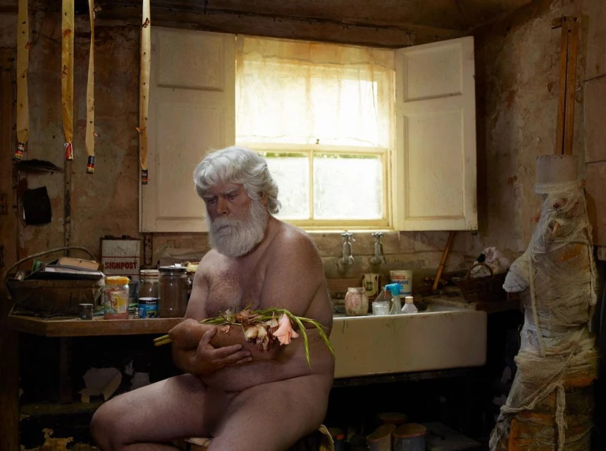 An older adult male with a beard sits shirtless in a rustic kitchen, cradling flowers, with natural light from a window.