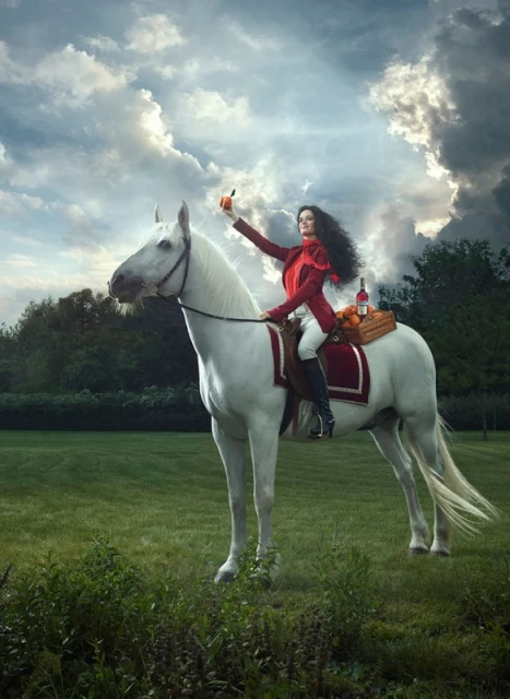 Woman in red jacket riding a white horse in a grassy field with cloudy skies, holding up a piece of fruit and carrying oranges and wine.