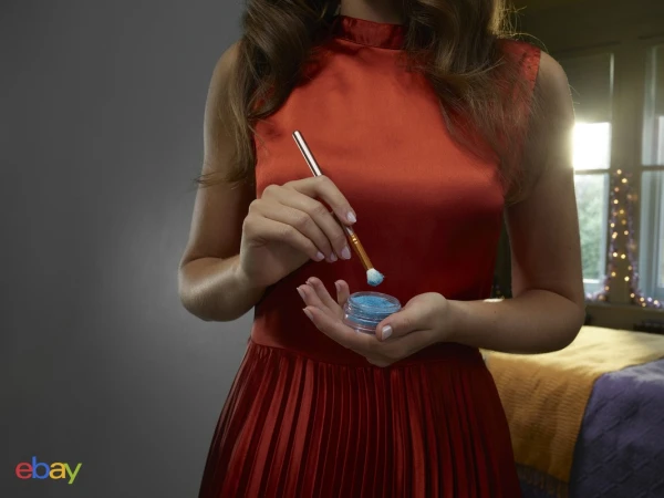 Woman in red dress applies blue eyeshadow with brush. Cosmetic jar in hand. Backlit indoor scene. Beauty and fashion concept.