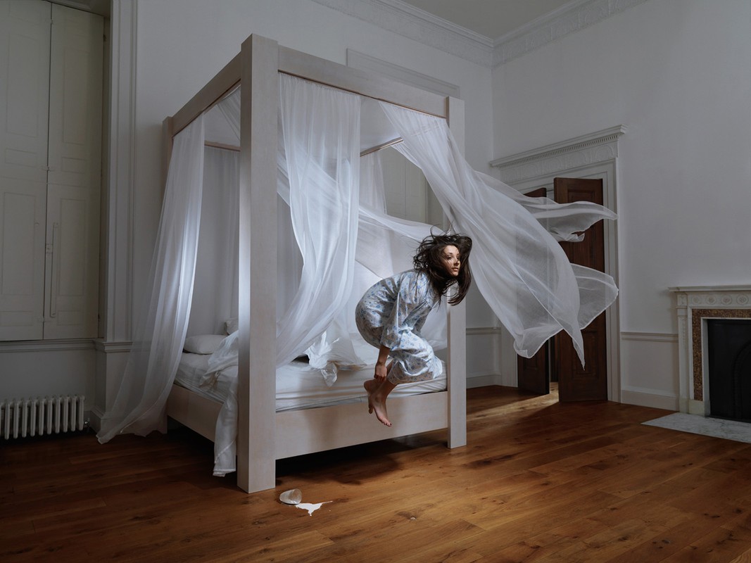 Young woman in pajamas leaps from a four-poster bed with flowing white curtains in a white-walled room with hardwood floors and a fireplace.