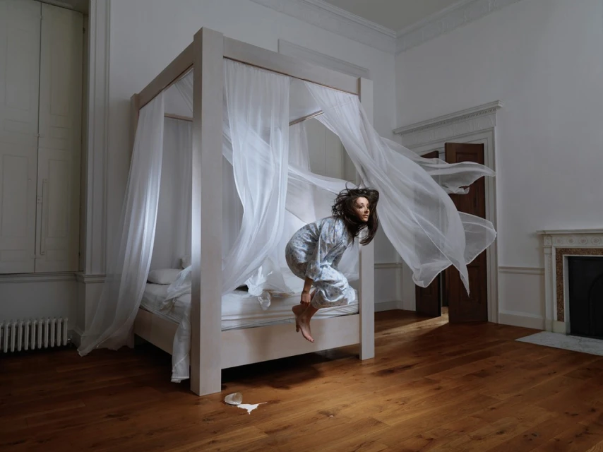 Young woman in pajamas leaps from a four-poster bed with flowing white curtains in a white-walled room with hardwood floors and a fireplace.