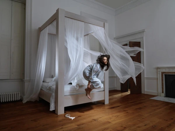 Young woman in pajamas leaps from a four-poster bed with flowing white curtains in a white-walled room with hardwood floors and a fireplace.