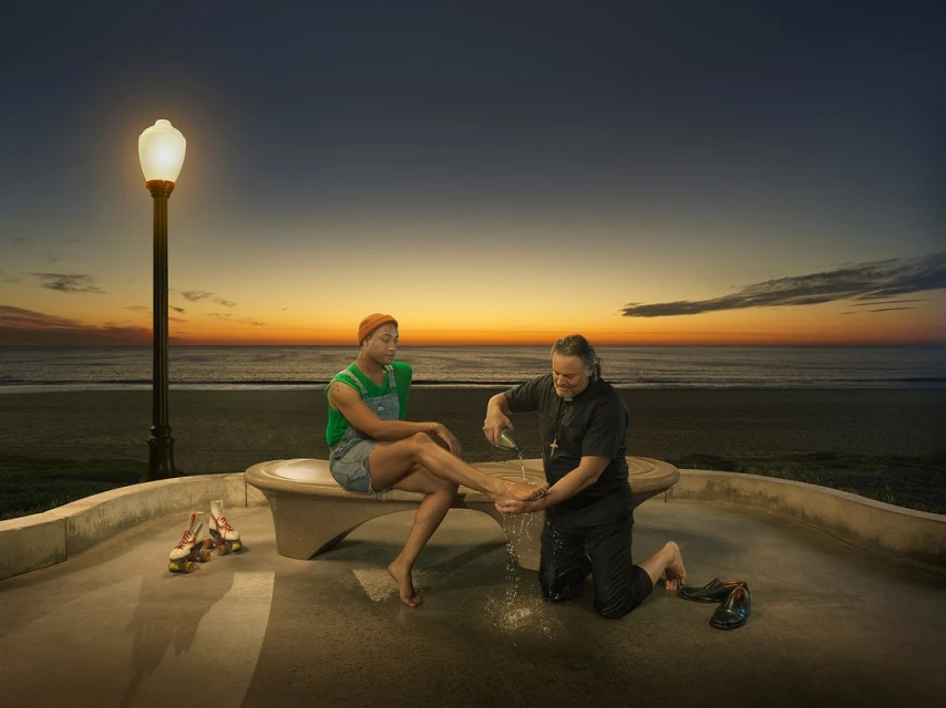 A man in a priest's collar washes the foot of a person sitting on a bench next to roller skates at the beach at sunset.