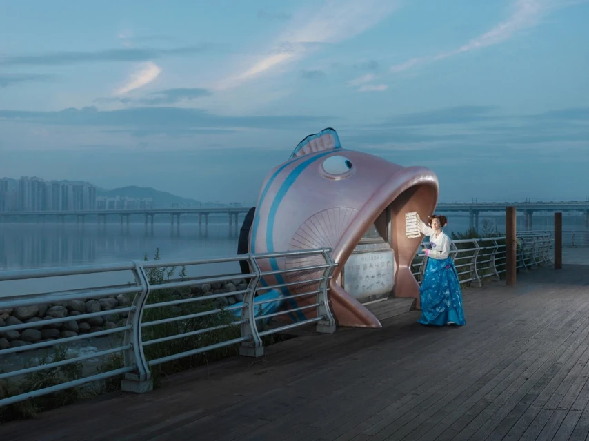 Woman in traditional Korean dress interacts with a large fish sculpture on a boardwalk with city buildings in the background.