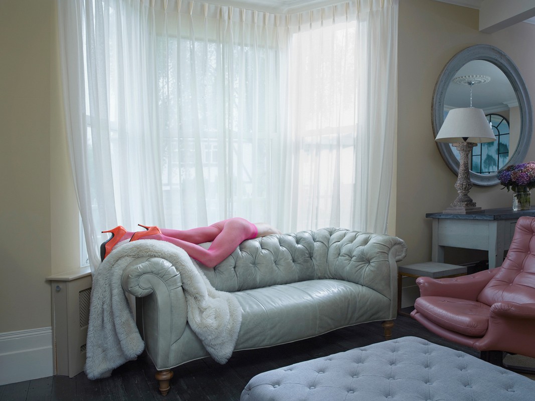 Interior view of a living room featuring a woman lying on a gray velvet chaise lounge with orange heels, sheer curtains and furniture.