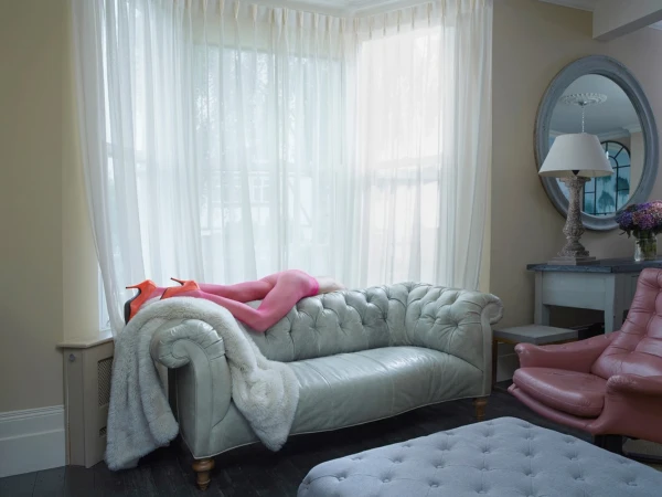 Interior view of a living room featuring a woman lying on a gray velvet chaise lounge with orange heels, sheer curtains and furniture.