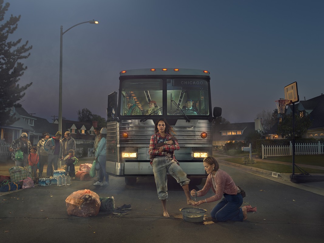 Street scene at night with a bus, woman standing with injured foot, another woman kneeling to help, and a group of people nearby.