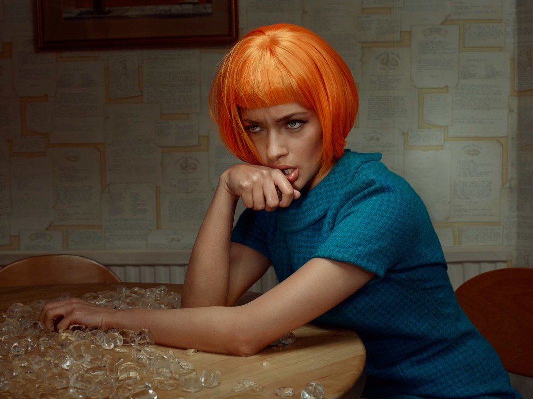 Young woman with orange wig poses at a table covered with ice against a wallpapered wall, creating an edgy and captivating scene.
