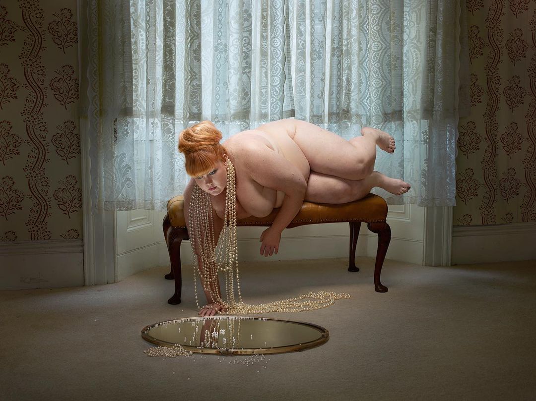 Overweight woman laying on upholstered bench in front of a mirror covered in pearl necklaces in an ornate interior room with wallpaper.