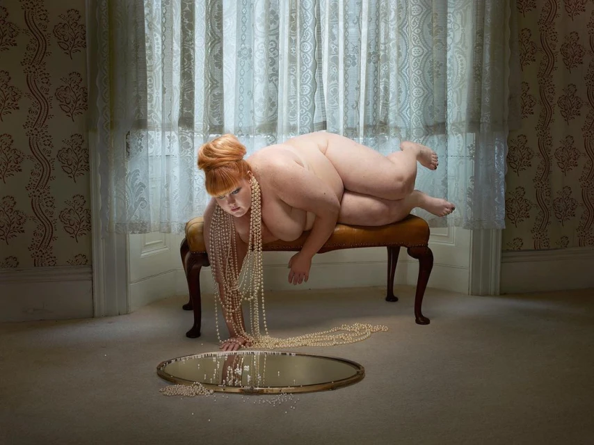 Overweight woman laying on upholstered bench in front of a mirror covered in pearl necklaces in an ornate interior room with wallpaper.