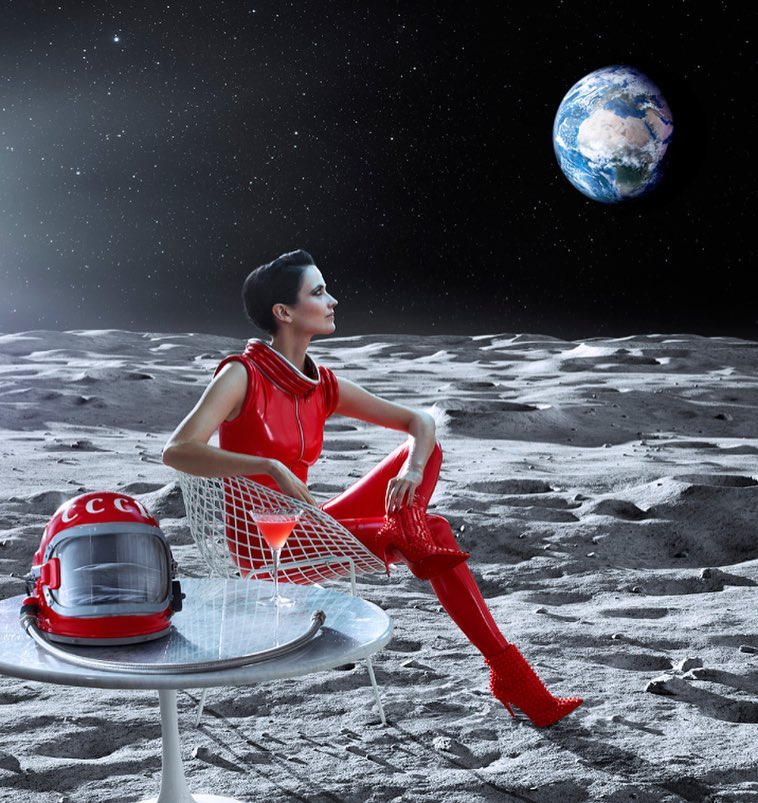 Woman in red spacesuit sits on the moon near a table with a cocktail, helmet, and Earth in the sky in a stylized, space-themed editorial.