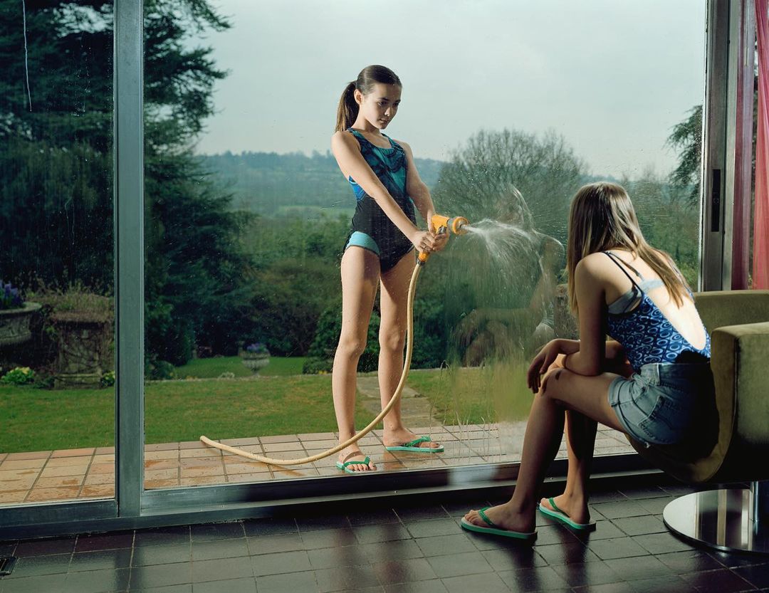 Two teenage girls in swimwear outdoors; one sprays water with a hose, the other sits near the window with a view of the greenery beyond.