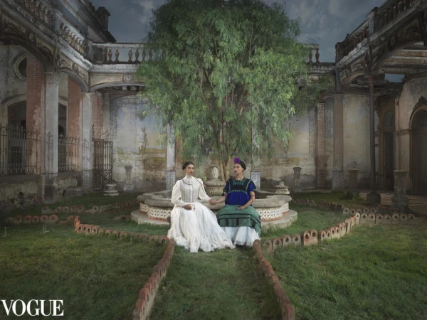 Two women in traditional garments seated near a fountain inside a decaying historic courtyard with a large tree