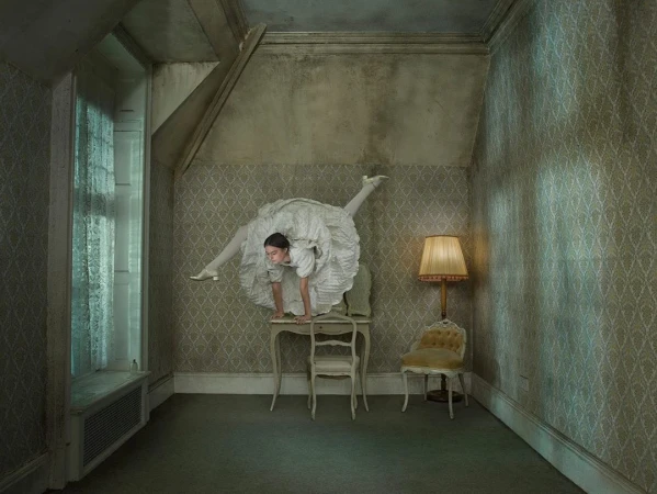 A woman in a large white dress poses in a room performing a handstand on a small table with a lamp and chair to the side.