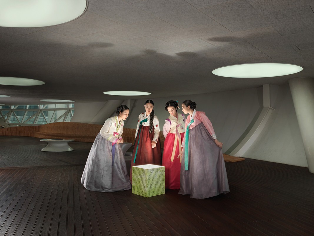 Four women in hanboks stand in modern interior around a glowing cube, lit by overhead lights, with geometric architectural features.