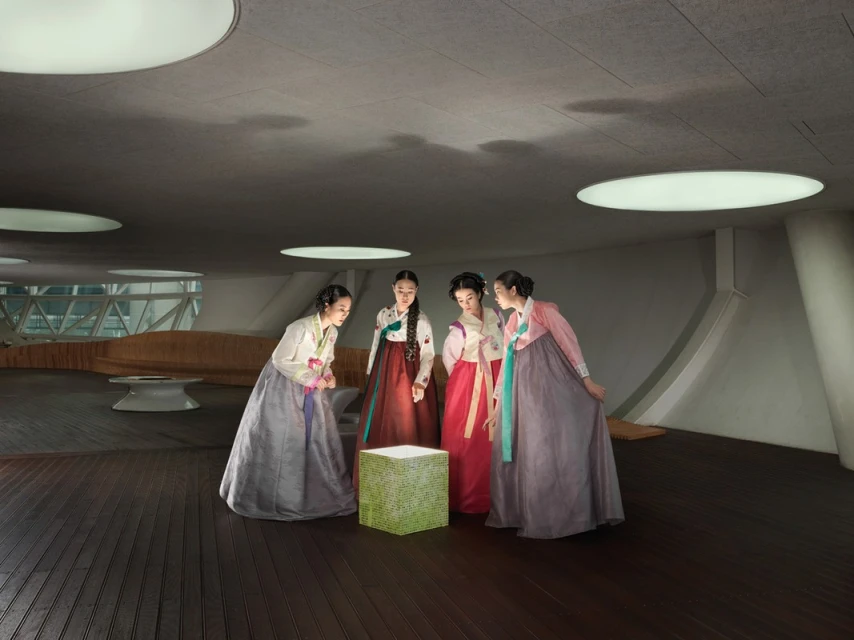 Four women in hanboks stand in modern interior around a glowing cube, lit by overhead lights, with geometric architectural features.