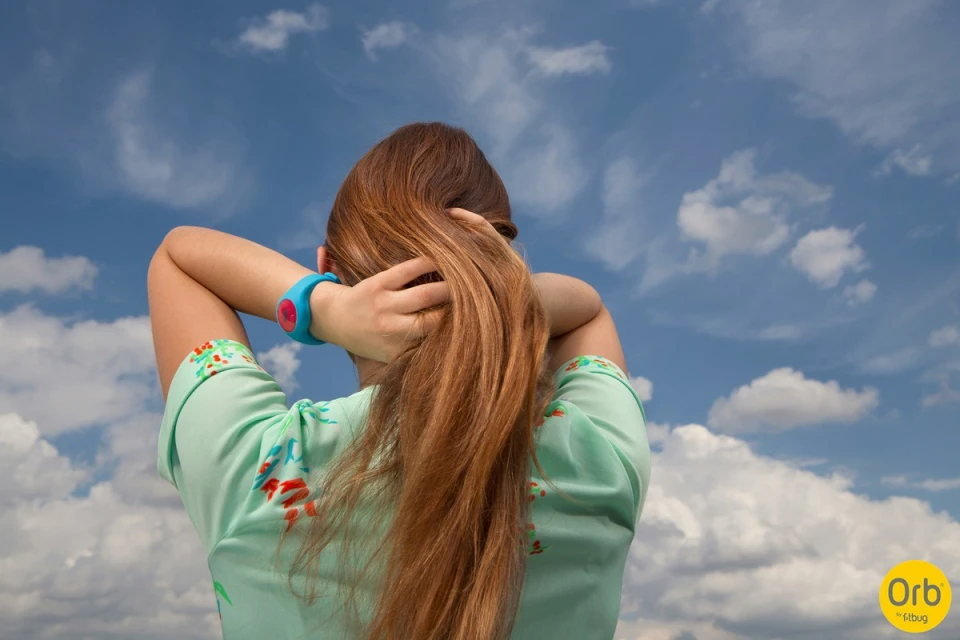 Back view of a person with long brown hair putting their hair up against a blue sky with clouds, wearing a light green top.