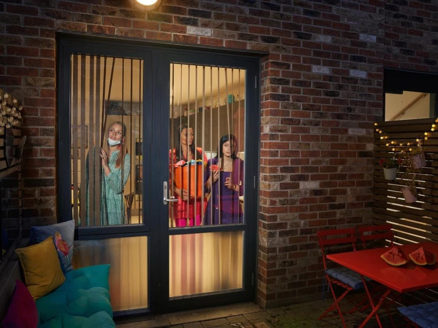 Three women visible through the windowed door in a brick house. One woman holds a container, another a drink, and the third peeks.