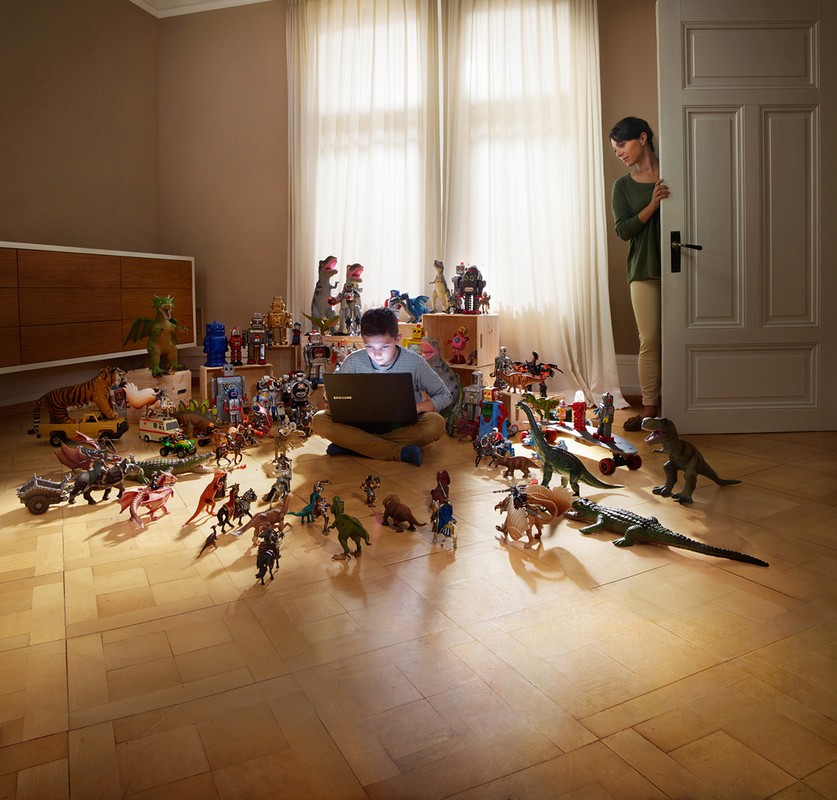 Boy sits cross-legged with laptop amid toy robots and dinosaurs in a light-filled room as a woman peeks from behind a door.