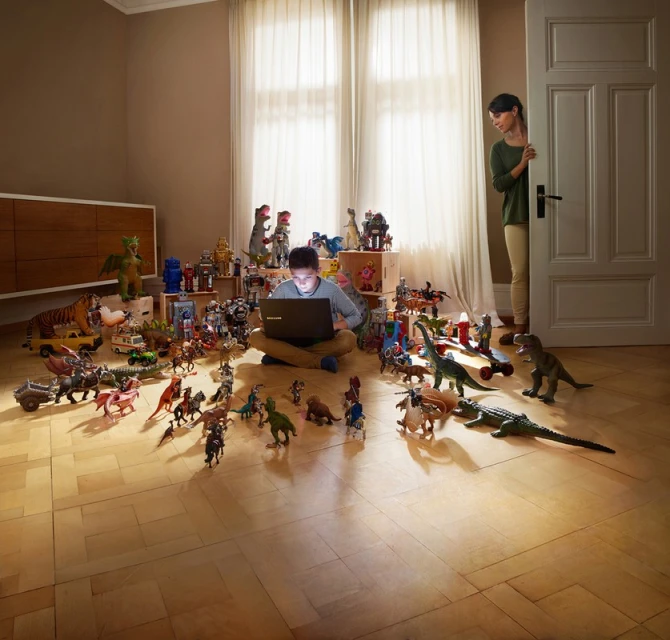 Boy sits cross-legged with laptop amid toy robots and dinosaurs in a light-filled room as a woman peeks from behind a door.