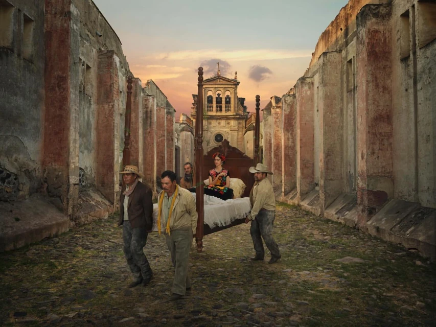 Frida Kahlo on a bed carried by four men through ruined city streets, set against a building and dusk sky.
