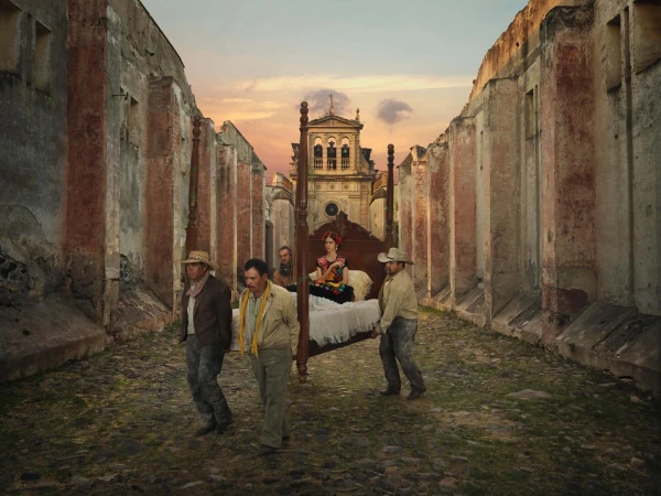 Frida Kahlo on a bed carried by four men through ruined city streets, set against a building and dusk sky.