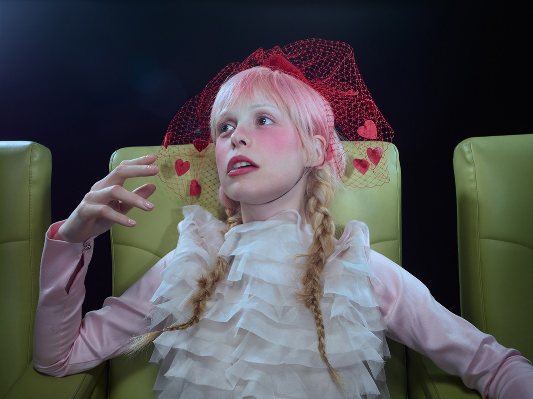 A model with pink hair and a red heart-themed veil is sitting in a green chair, looking to the side with a slight expression in a studio setting.