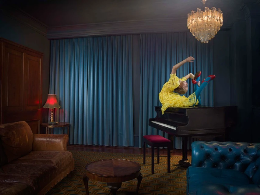 A woman in a yellow dress sits on a piano in an ornate room with blue walls and a chandelier in a visually striking composition.