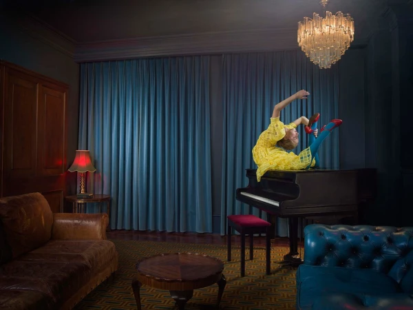 A woman in a yellow dress sits on a piano in an ornate room with blue walls and a chandelier in a visually striking composition.