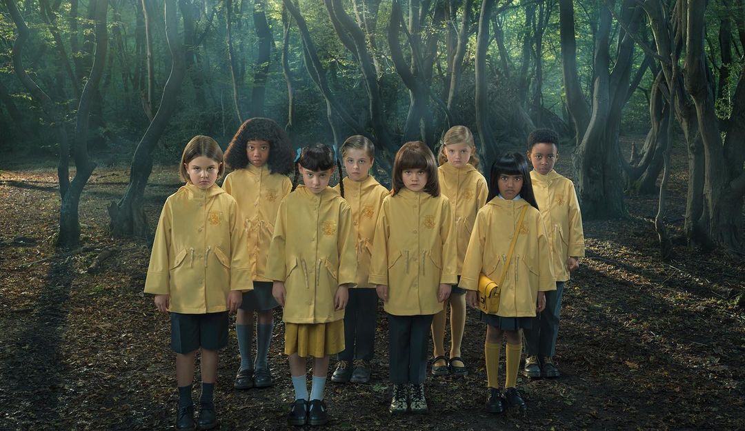 Group of children in yellow raincoats stand in a forest, looking directly at the camera with serious expressions, captured in eye-level shot.