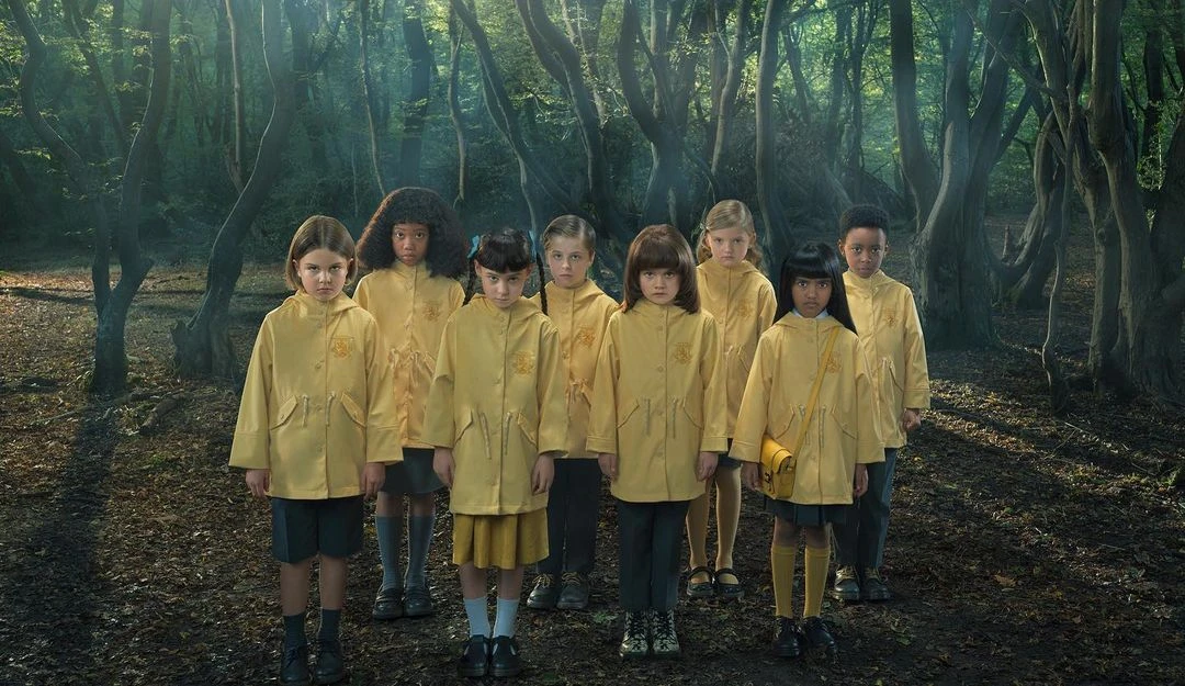 Group of children in yellow raincoats stand in a forest, looking directly at the camera with serious expressions, captured in eye-level shot.
