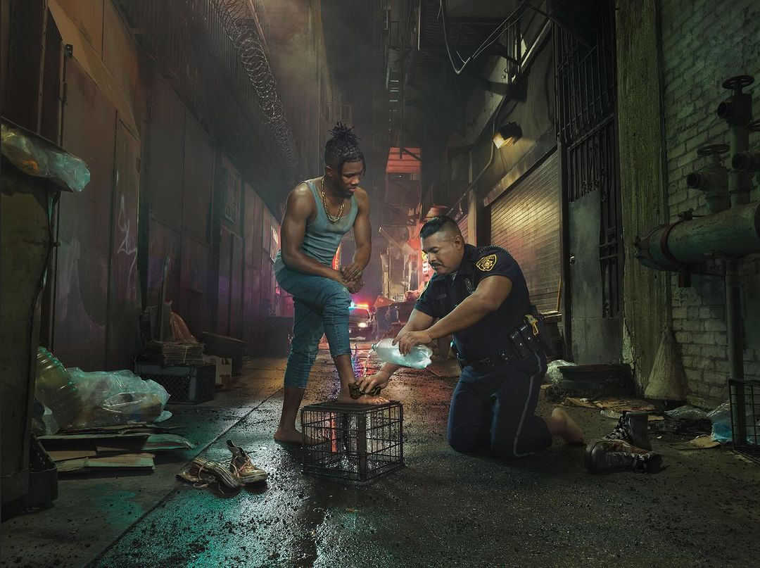 A barefoot man stands on a metal cage as a police officer pours water on his foot in an alley at night, next to shoes and debris.