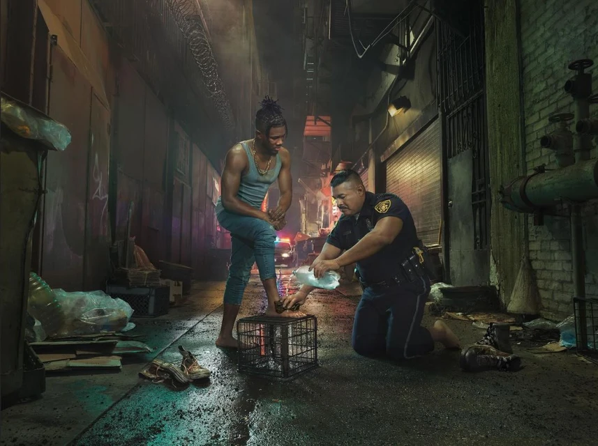 A barefoot man stands on a metal cage as a police officer pours water on his foot in an alley at night, next to shoes and debris.