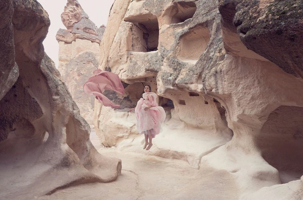 Woman in flowing pink gown appears to float amid rock formations in Cappadocia, Turkey. Dreamy fashion photography.