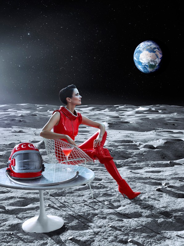 Woman in red space suit sitting on the Moon with the Earth in view. She is at a table with a drink next to a Soviet space helmet.