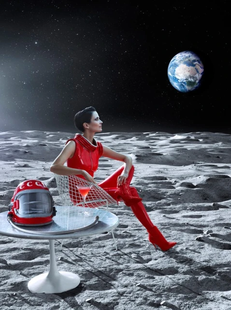 Woman in red space suit sitting on the Moon with the Earth in view. She is at a table with a drink next to a Soviet space helmet.