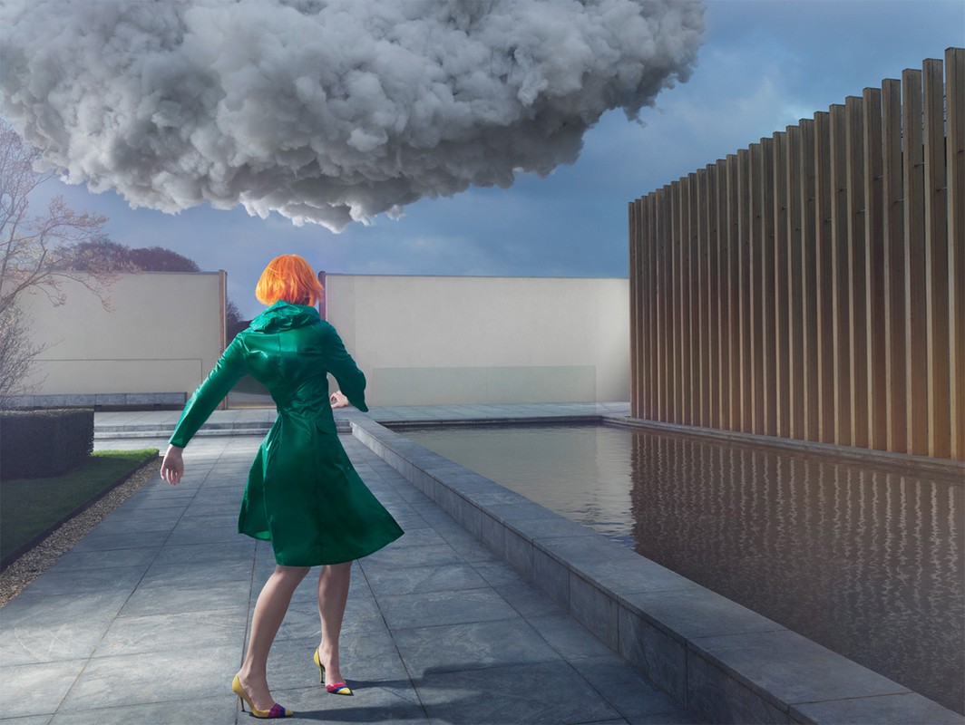 A woman with orange hair wearing a green coat stands near a pool with a large cloud overhead and a wood slat wall.