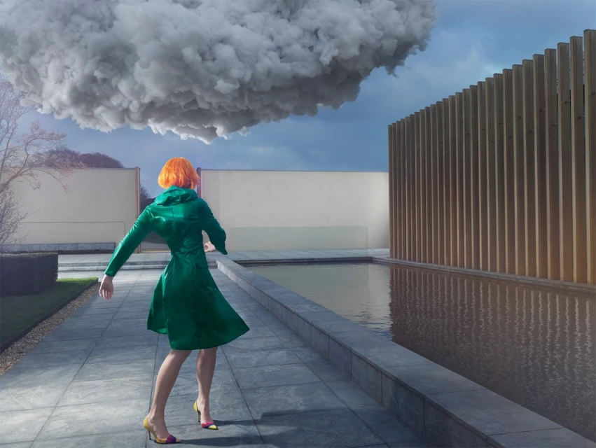 A woman with orange hair wearing a green coat stands near a pool with a large cloud overhead and a wood slat wall.
