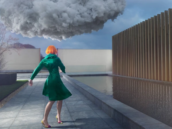A woman with orange hair wearing a green coat stands near a pool with a large cloud overhead and a wood slat wall.