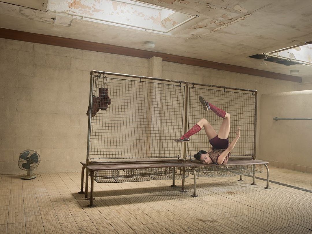 Woman in vintage sportswear performing floor exercises in a gritty indoor location, with boxing gloves and an old fan visible.
