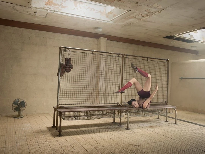Woman in vintage sportswear performing floor exercises in a gritty indoor location, with boxing gloves and an old fan visible.