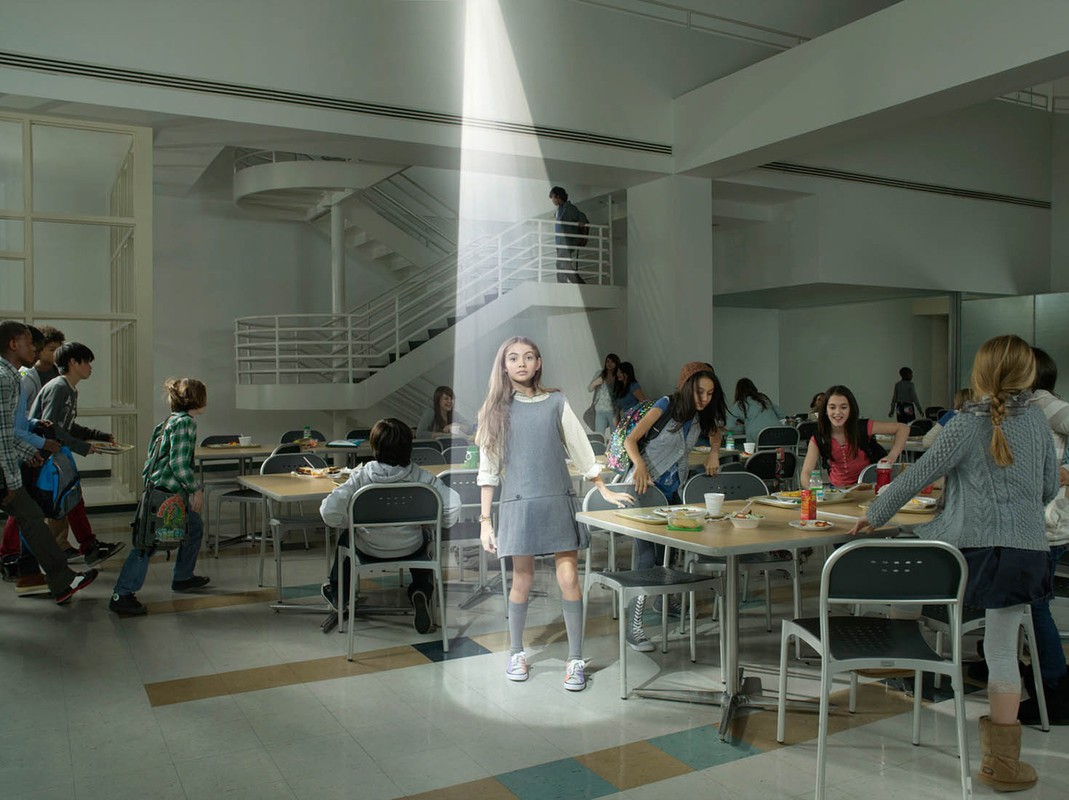 A young girl stands under a beam of light in a school cafeteria, surrounded by other students and tables, creating a surreal scene.