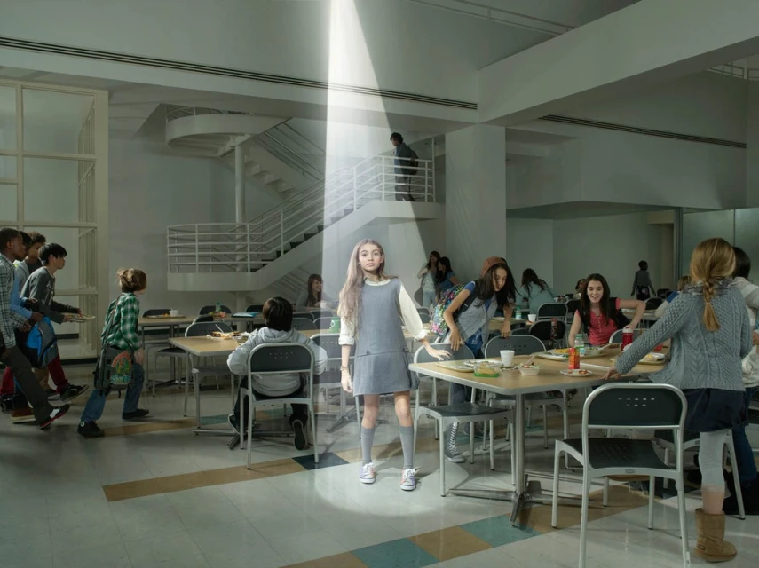 A young girl stands under a beam of light in a school cafeteria, surrounded by other students and tables, creating a surreal scene.